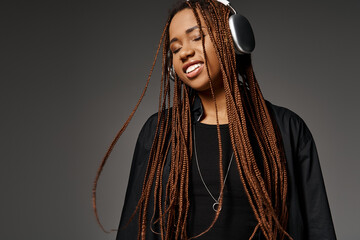 pleased and young African American woman in 20s with headphones listening to music on grey backdrop