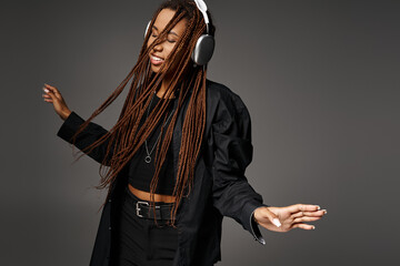 joyful and young African American woman in 20s with headphones dancing to music on grey backdrop
