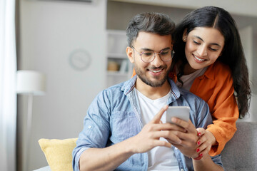 Cheerful indian couple using cellphone, shopping online, spouses sitting on sofa at home interior
