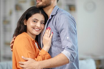 Affectionate hidnu couple hugging while standing in living room interior, free space. Romantic...