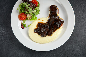 Braised beef cheeks over smooth mashed potatoes, garnished with fresh salad, top view. A comforting...