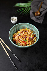 Chinese fried noodles with chicken and vegetables in ceramic bowl on black concrete background....