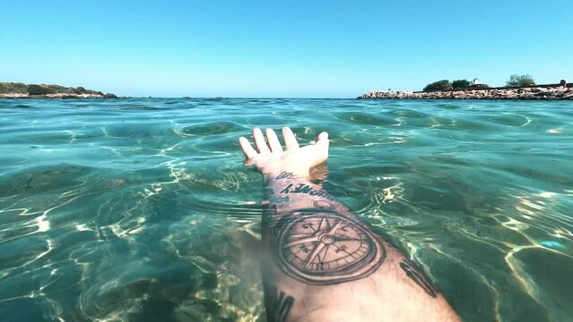 Pov of arm hand man with compass traveler tattoo enjoying blue crystal transparent tropical sea ocean water. One adult male people enjoy beach lifestyle alone. Outdoor swimming leisure activity