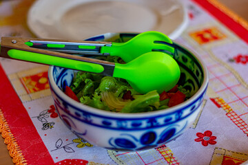 tigela azul e branca com salada de alface e tomate com um colher verde, ao fundo um prato branco - obrazy, fototapety, plakaty