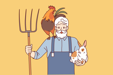 Elderly man farmer engaged in agriculture and livestock raising holds rake and rabbit with rooster. Happy grandfather farmer rejoices at having own farm and possibility of country life