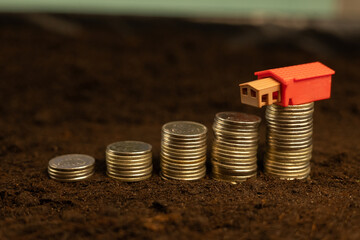 Money on land with house. Stacks of coins in ascending order are on the ground and on top of them is a small house. Mortgage concept for the purchase of land and house.
