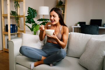 Beautiful young woman is sitting on the sofa in the living room and eating oatmeal