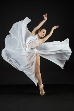 A ballerina in a white dress and pointe shoes stands on her toes and dances on a black background