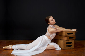 Ballerina in a white dress and pointe shoes sits on the floor on a black background