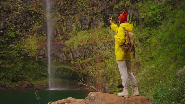 A girl traveler in a yellow jacket and a backpack reached a beautiful, inaccessible waterfall and took pictures of it on her phone