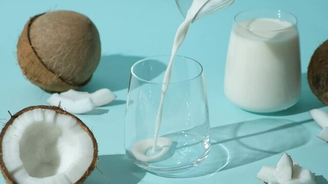 Coconut milk pouring into a glass among coconuts on a blue background, slow motion.
