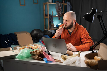 Bearded man eating pizza and working in messy, cluttered freelancer room with piles of clothes