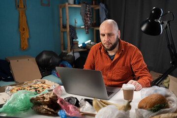 Busy male freelancer using laptop surrounded by tons of garbage and food in his apartment