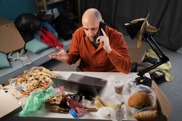 Slatternly bearded man sitting before laptop and holding smartphone among leftovers, food, bags and...