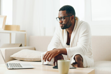 Smiling African American Freelancer Typing on Laptop in a Modern Home Office African American man in bathrobe sitting on a sofa in his living room, happily working on his laptop in the morning The