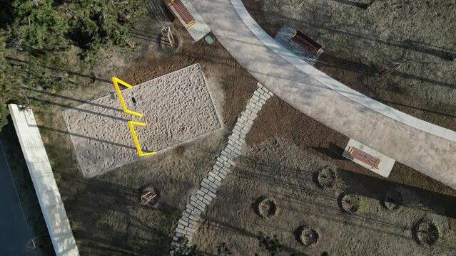 a circle-shaped wood bench with a tree growing in the middle. swings and sandy paths in the park. a bed of flowering bulbs. yellow gates and pine trees looking vertically down, drinking fountain