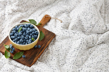 Yellow bowl of fresh blueberries on wooden board on bed blanket
