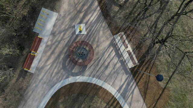 a circle-shaped wood bench with a tree growing in the middle. swings and sandy paths in the park. a bed of flowering bulbs. yellow gates and pine trees looking vertically down, drinking fountain