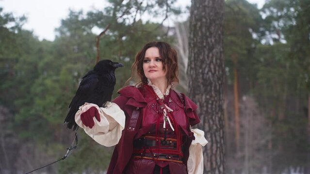 A woman in a red dress is holding a black bird on her shoulder in the forest