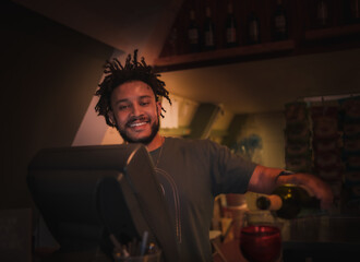 Young African American barman poring red wine in glass and smiling in dimly lit restairamt