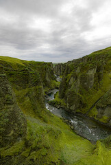 River flows through a canyon with towering mountains on each side. Watercourse cuts through the natural landscape, surrounded by lush grass in the valley, Fjadrargljufur Canyon