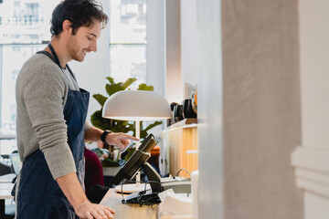 Side view portrait of young good-looking caucasian barista bartender barman shop assistant vendor owner working on the cash point in cafeteria restaurant coffee shop