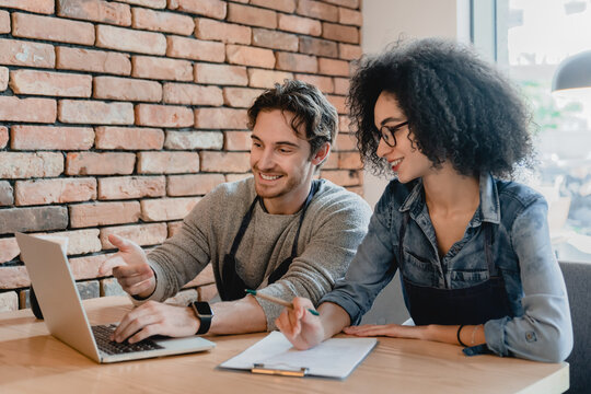 Two cafeteria staff working on laptop and writing down together in loft cafeteria. Educational promotional training for staff, e-learning for shop assistants in restaurant. New trainees