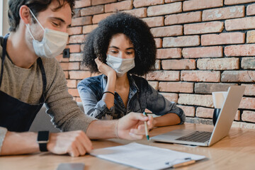 Two cafe workers wearing medical masks against Covid19 working on laptop in loft cafeteria. Multicultural cafe staff vendors shop owners counting budget, creating development plan