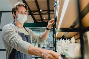 25s handsome waiter in medical mask against coronavirus taking glasses from the shelf in coffee...