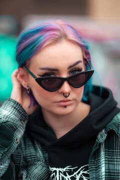 Young woman with colorfull blue hair, piercings and sunglasses posing. Wearing flannel and hoodie. Urban background.