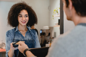 Smiling attractive afro waitress servicing the customer using swiping machine in cafeteria. E-banking e-commerce. Payment with credit card debit account. Selling buying food and drinks