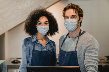 Two young cafe workers standing at cash point wearing medical masks against covid 19. Social...
