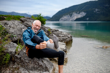 Active elderly man putting on sock after barefoot walk in cold water in mountain lake. Senior...