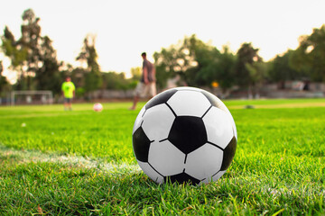 Imagen horizontal de un balón de futbol Soccer color blanco con negro y al fondo de la cancha de futbol desenfócanos padre e hijo jugando  - obrazy, fototapety, plakaty