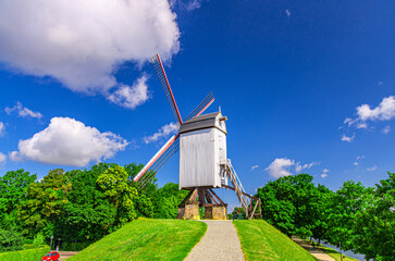 Traditional old windmill on green hill in Brugge city, blue sky white clouds background in summer sunny day, typical white wind mill with sails, path in park with green trees, Flemish Region, Belgium