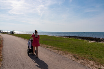 Loving mother walking with baby stroller along coastal road in Medulin, Istria peninsula, Croatia, Europe. Idyllic coastline of Kvarner Gulf in Adriatic Mediterranean Sea. Travel destination in summer