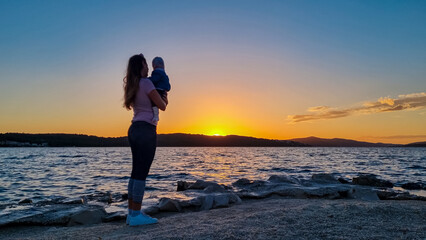Silhouette of loving mother holding small toddler at romantic sunset in Okrug Gornij, Split-Dalmatia, Croatia. Coastline of island Otok Ciovo, Adriatic Sea in summer. Family travel vacation concept