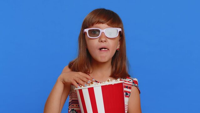 Excited young cute school girl in 3D glasses eating popcorn, watching interesting tv serial, sport game, film, online social media movie content. Teen female child kid on studio blue background