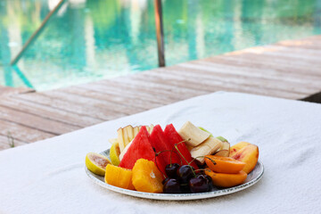 Plate with fresh fruits on sun lounger near outdoor swimming pool. Luxury resort