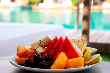 Plate with fresh fruits on table near sun lounger. Luxury resort with outdoor swimming pool
