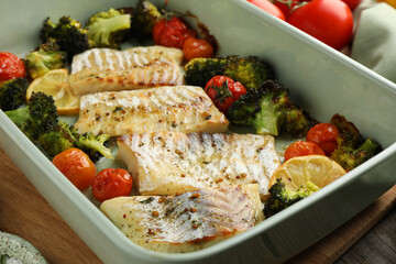 Pieces of delicious baked cod with vegetables, lemon and spices in dish on table, closeup