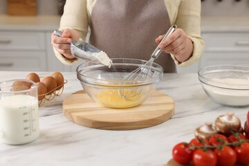Woman whisking eggs while adding flour into bowl at light marble table indoors, closeup