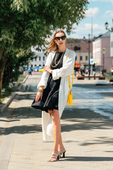 Stylish woman in a black dress and a white cardigan with a bright yellow handbag and a gold chain. A summer dress with frills flutters in the wind. Full length girl in the city in summer.