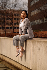 A young woman is casually perched on a concrete ledge, dressed in a stylish grey blazer, striped trousers, and pink sneakers, enjoying a sunny day in an urban setting.