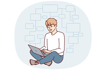 Young man programmer sits cross-legged on floor with laptop on lap and creates web application. Guy IT developer thinks over flowchart modern website or blockchain project