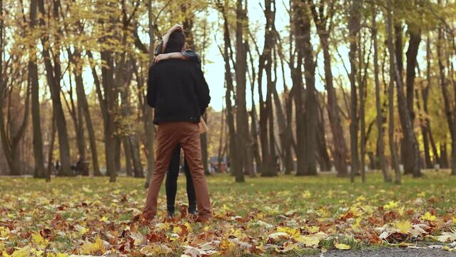 Young loving couple walking outside in the city park in sunny weather, dancing on orange yellow red maple leaves, hugging smiling kissing laughing spending time together. Autumn, fall season 
