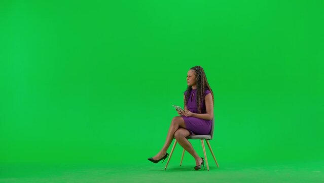 Female in dress isolated on chroma key green screen background. Full shot african american woman tv news host sitting holding tablet and taking interview.
