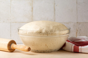 Glass bowl with fresh homemade yeast dough on a kitchen table with utensils.