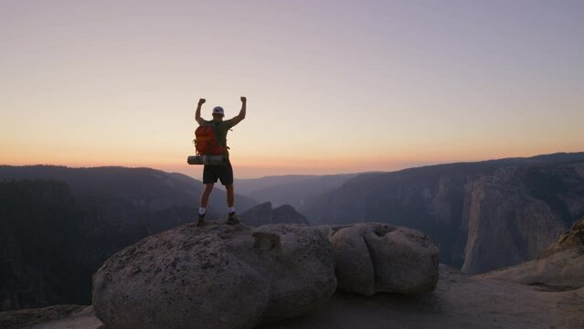 Amazing view of dark mountain range with orange skyline, happy male hiker on a stone on a summit raising hands up with delight, overwhelmed with emotions. High quality 4k footage