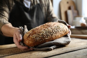 Man holding loaf of fresh bread at wooden table indoors, closeup